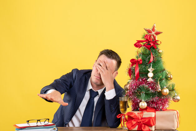 沮丧的男人前视图沮丧的男子坐在桌旁 用手捂住眼睛靠近圣诞树和黄色的礼物坐着商人男人