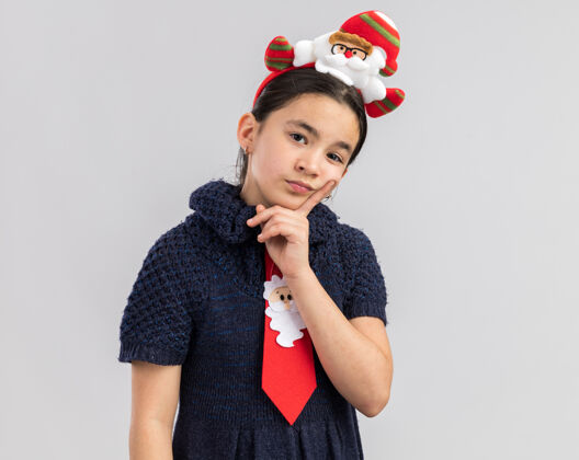 搞笑穿着针织连衣裙的小女孩头上戴着一条红色领带 上面戴着一个滑稽的圣诞边 看上去很困惑红快乐圈