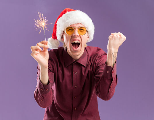 金发快乐的年轻金发男子戴着圣诞帽 戴着圣诞眼镜 拿着节日火花灯 在紫色的墙上做着“是”的手势快乐闪闪发光圣诞老人