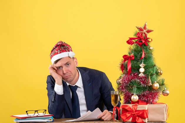 男人一个戴着圣诞帽的沮丧男人坐在圣诞树旁的桌子前 黄色的礼物帽子黄色商人