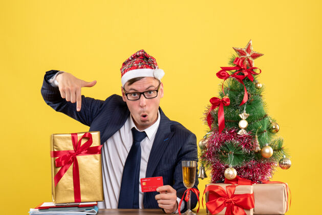 商务男士前视图中的商人指着他坐在圣诞树旁的桌子上的礼物和黄色的礼物黄色圣诞西装
