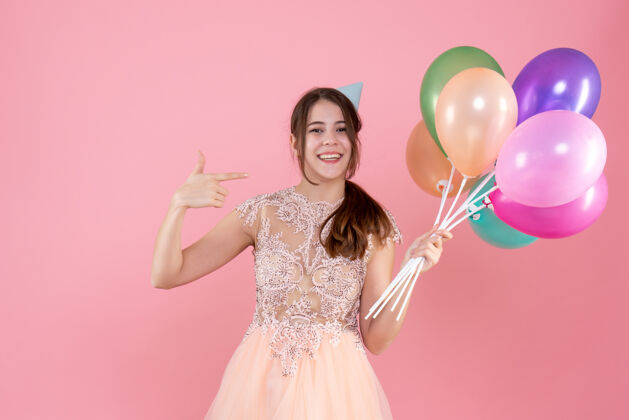 手持快乐的派对女孩 戴着派对帽 拿着气球 手指指着粉色的自己肖像可爱粉色
