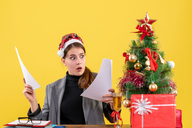 工作正面图戴着圣诞帽的大眼睛女孩坐在桌子旁看着圣诞树和礼物鸡尾酒文件人坐着