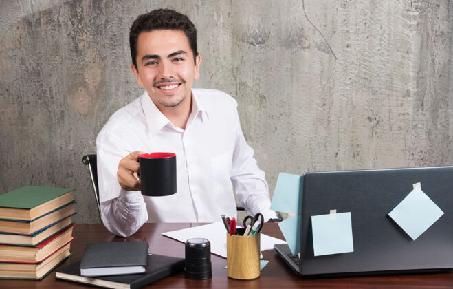 办公室年轻的商人在办公桌前端着一杯茶人拿着微笑