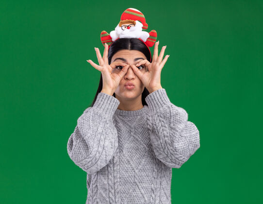 年轻人令人印象深刻的年轻白人女孩戴着圣诞老人的头带做看手势用手作为望远镜隔离在绿色的墙上圣诞老人表情圣诞节