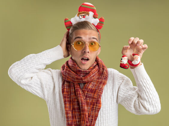 围巾令人印象深刻的年轻帅哥戴着圣诞老人的头带和围巾拿着圣诞老人的圣诞饰品 手放在脑后 隔离在橄榄绿的墙上戴橄榄年轻