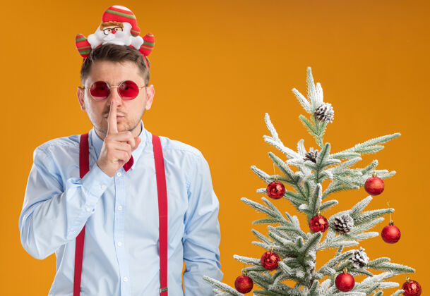手指戴吊带领结的年轻人站在圣诞树旁 戴着圣诞老人和红眼镜 手指放在嘴唇上 在橙色的墙上做着沉默的手势年轻人站立制作