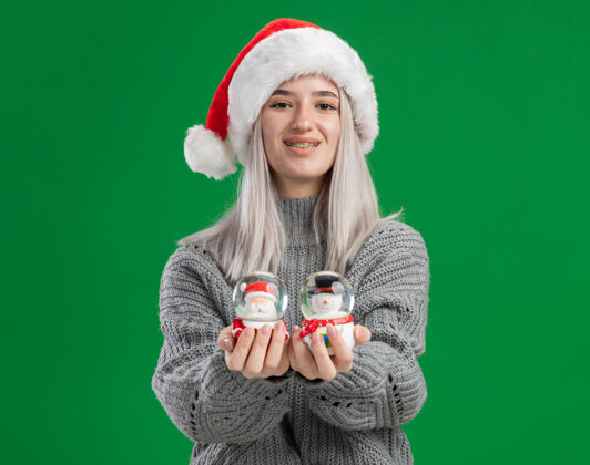 圣诞节穿着冬季毛衣 戴着圣诞帽 拿着圣诞玩具雪球的快乐金发女郎站在绿色背景下 看着相机 开心地微笑着抱着金发圣诞老人