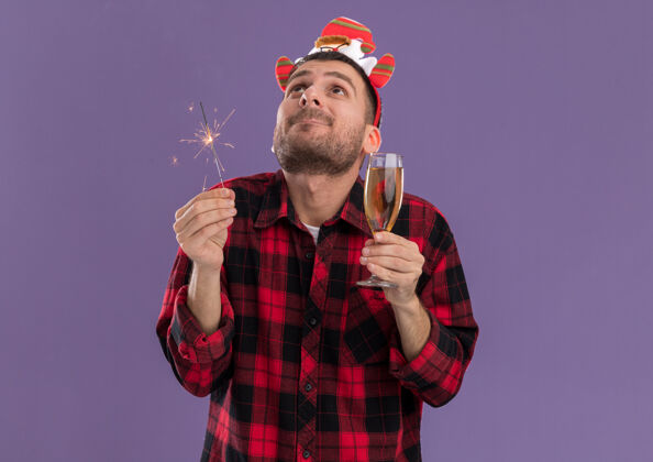 香槟令人印象深刻的年轻白种人戴着圣诞老人的头带 拿着节日的火花和香槟杯 在紫色的墙上看着孤立的复制空间圣诞老人闪亮头带