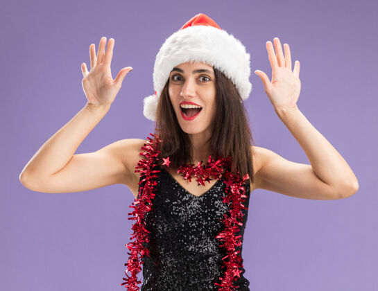 女孩惊讶的年轻漂亮女孩戴着圣诞帽 脖子上戴着花环 在紫色背景上举手孤立圣诞节提高手