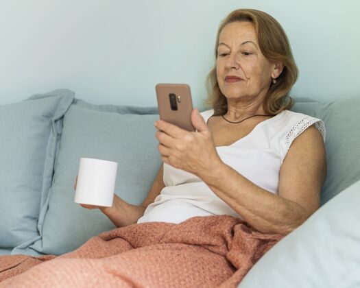 防护大流行期间 家里的老太太在喝咖啡和使用智能手机杯子冠状病毒Ncov