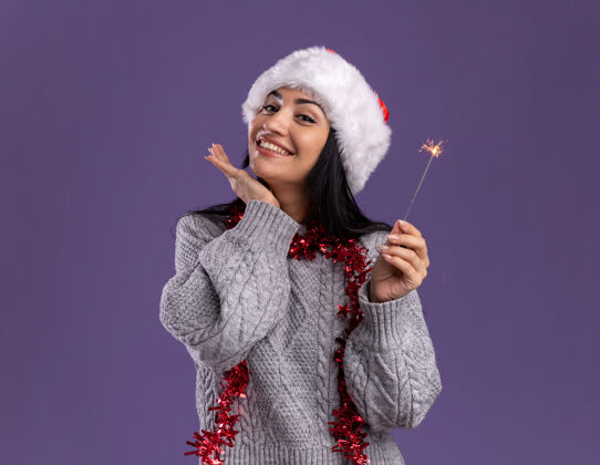 下巴微笑着的年轻白人女孩戴着圣诞帽 脖子上戴着金箔花环 手里拿着节日火花灯 看着相机 手放在下巴下面 紫色背景上 留着复印空间帽子周围微笑