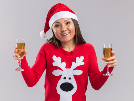 微笑微笑的亚洲女孩戴着圣诞帽 穿着毛衣 手里拿着一杯香槟 背景是白色的杯子亚洲人香槟