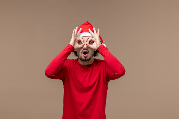 喜剧演员前视图年轻的男性透过手指看棕色背景的情感圣诞假期封面衣服人
