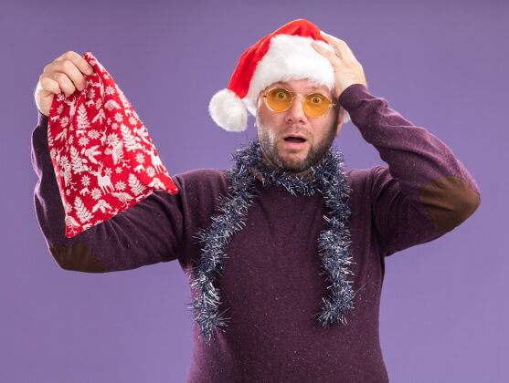 礼物一个戴着圣诞帽 脖子上戴着金箔花环 戴着眼镜 手拿圣诞礼物袋 看着摄像机 手放在头上 在紫色背景下与世隔绝围着手捧圣诞老人
