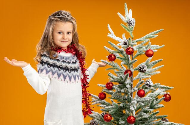 高兴可爱的小女孩站在圣诞树旁 戴着头饰 脖子上戴着花环 两手分开放在橙色的背景上圣诞快乐女孩手