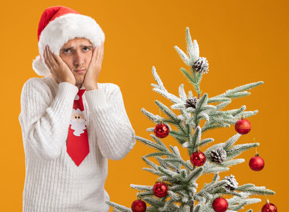 小伙子戴着圣诞帽 系着圣诞老人领带的年轻帅哥站在装饰好的圣诞树旁 手放在脸上看着隔离在橙色背景下的相机英俊看悲伤