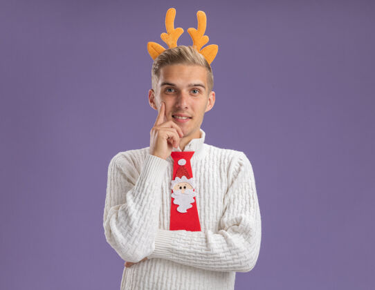 帅气高兴的年轻帅哥戴着驯鹿鹿角头带和圣诞老人领带看着相机触摸下巴孤立的紫色背景与复制空间小伙子圣诞老人头带