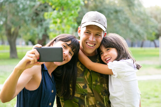智能手机快乐的军人 他的妻子和小女儿在城市公园里用手机自拍正面视图家庭团聚或回家的概念公园白种人肖像