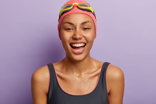 泳衣快乐女运动员专业游泳运动员在游泳池训练后全身湿透 穿着泳衣 戴帽子和护目镜 露出洁白的牙齿 隔离在紫色的墙壁上夏季时间和积极的休息理念快乐运动健身