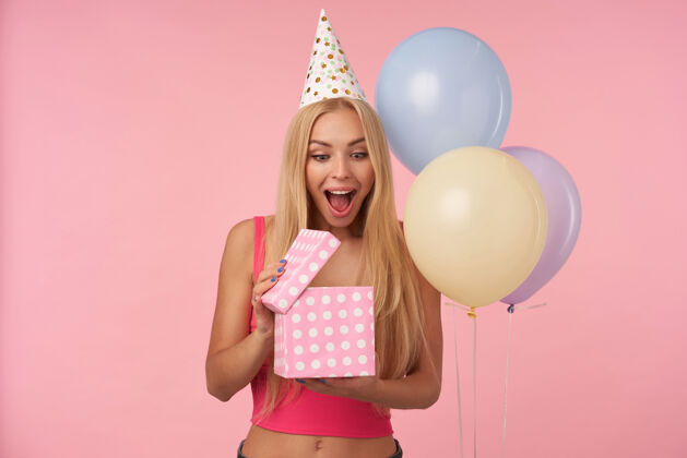 漂亮快乐的金发女郎 留着长长的头发 打开礼物 对自己的内容感到惊讶 在粉色背景下摆出五颜六色的气球 睁大眼睛和嘴巴五彩20多岁休闲