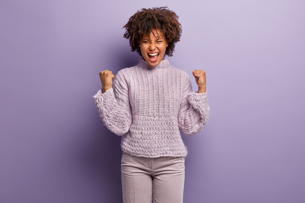 高兴快乐的黑皮肤女模特画像与凯旋握拳 有着喜出望外的表情 表示收到凯旋后的幸福 穿着紫色衣服 在室内积极手势快乐毛衣非洲