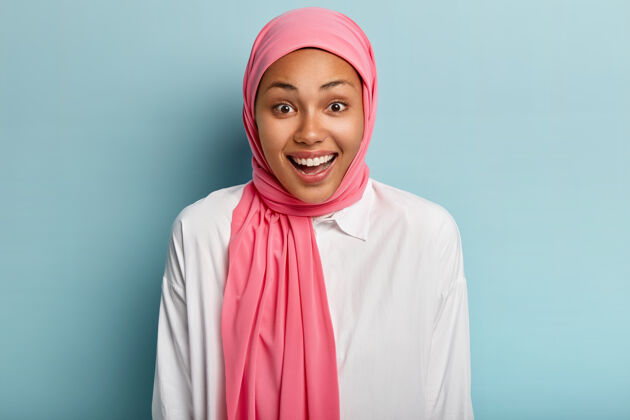 信仰喜怒无常的妇女欢笑 有愉快的表情 表达真挚的感情 戴着粉红色的头饰 露出白色的牙齿 隔着蓝色的墙壁人类情感的概念过度运动凝视头巾