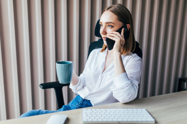 電子商務在咖啡杯旁的辦公桌上 一位可愛的年輕女士一邊用手提電腦一邊打電話女性現代辦公室