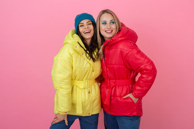 时尚两个迷人的女孩在粉红色的背景下 穿着鲜艳的红色和黄色的彩色冬季羽绒服摆姿势衣服微笑拥抱