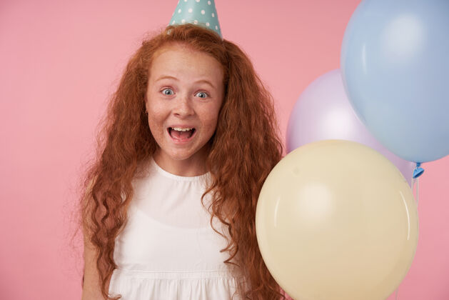 女孩喜气洋洋的红头发女孩的肖像 长卷发 白色连衣裙 戴着生日帽 为得到生日礼物而兴奋和惊讶 在粉色背景下愉快地看着相机孩子庆祝气球