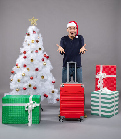 圣诞树在白色圣诞树旁 一个拿着红色手提箱的惊喜男人 身上挂着五颜六色的圣诞玩具购物者圣诞帽红色
