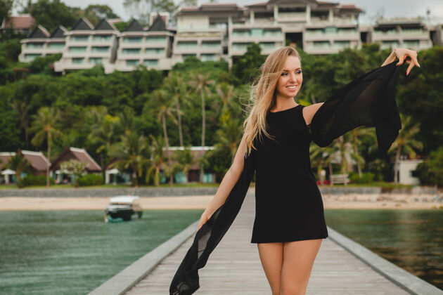 女人穿着黑色连衣裙的美丽迷人的女人在豪华度假酒店的码头上摆姿势 暑假 热带海滩摆姿势头发奢华