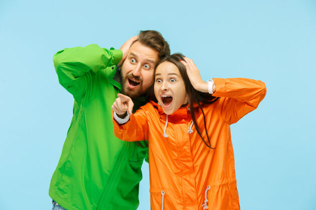 天气这对年轻的夫妇惊讶地指着左边 穿着蓝色的秋夹克在摄影棚摆姿势人类的负面情绪寒冷天气的概念女性和男性的时尚概念年轻酷外套
