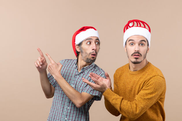 两个惊讶的家伙前视图两个戴着圣诞帽的惊讶的家伙在米色的孤立背景上展示了一些东西惊讶男人帽子
