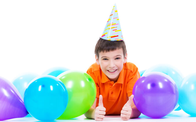 躺着穿着橙色t恤的快乐微笑男孩躺在地板上 手里拿着五颜六色的气球 竖起大拇指——孤立在白色的地板上竖起大拇指儿童男性