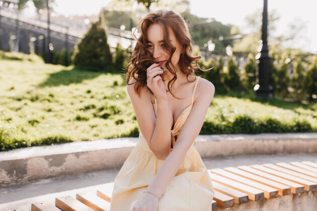 放松害羞的姜黄色卷发女孩坐在长凳上微笑着穿着优雅黄色连衣裙的漂亮女模特的户外照片时尚年轻微笑