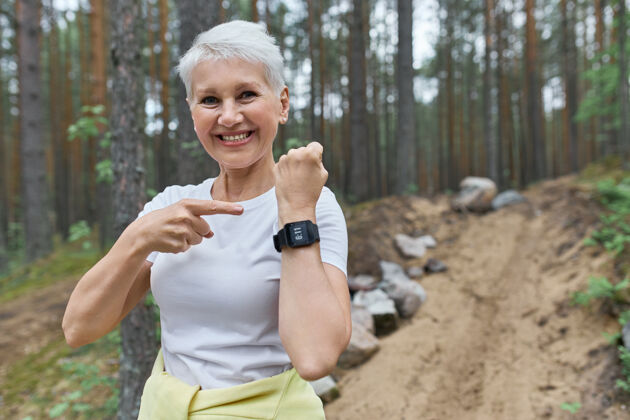 运动装快乐合身的退休女装运动衫微笑着宽大地指着展示的腕表智能手表手镯老年人户外