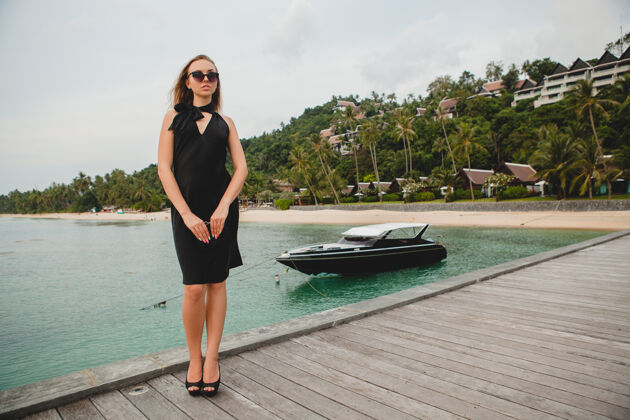 优雅奢华性感迷人的女人穿着黑色礼服在豪华度假酒店的码头上摆造型 戴着墨镜 暑假 热带海滩人热带旅游