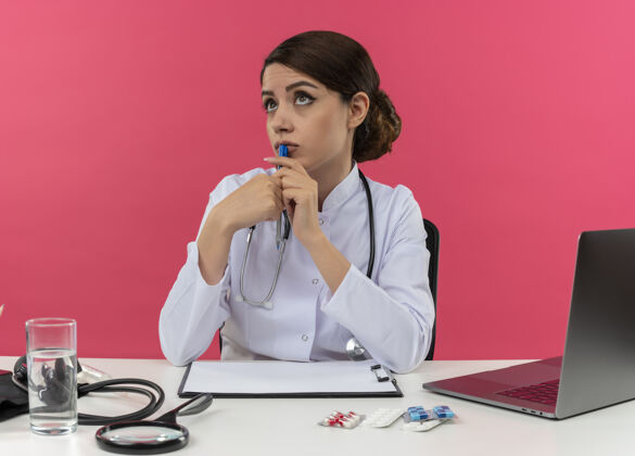穿想着年轻的女医生穿着医用长袍带听诊器坐在办公桌前拿着医疗工具拿着钢笔拿着复印空间在电脑上工作思考女医生