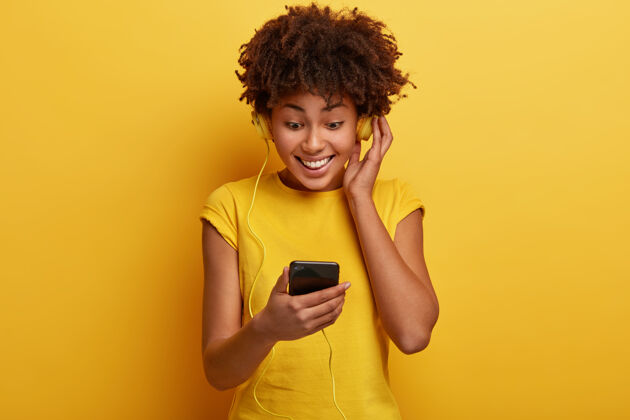 唱歌快乐的黑皮肤女人一边用耳机听悦耳的旋律 一边心情舒畅 专注在智能手机的屏幕上连接移动非洲