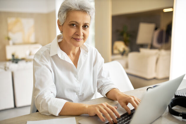 桌子一幅穿着白衬衫的50岁时尚开朗女作家的肖像画 她在工作中使用通用的电子设备 在她的新书中键入另一章计算机自信坐着的