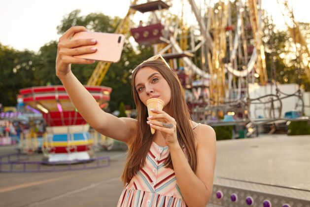 持有年轻漂亮的黑发女子站在游乐场的装饰物上 手里拿着智能手机 拿着冰激凌筒拍照阳光户外可爱