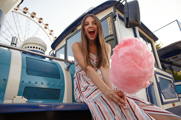 公园一头长发的快乐女子坐在游乐场上空的蒸汽火车上 手里拿着棉花糖 张大嘴巴 神采奕奕地看着坐着蒸汽年轻