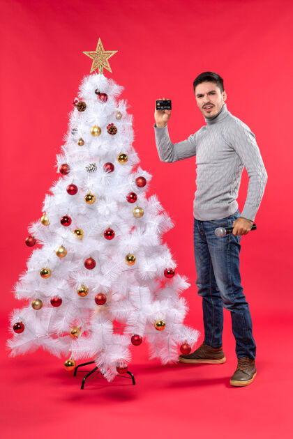 年轻圣诞气氛严肃的年轻人站在装饰好的圣诞树旁 拿着麦克风和电话自信地站着站着季节装饰
