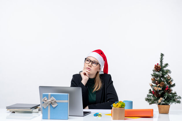 深思熟虑在办公室里 一个戴着圣诞老人帽子的漂亮女人坐在一张桌子旁沉思着 桌上放着圣诞树和一份礼物圣诞节女商人礼物