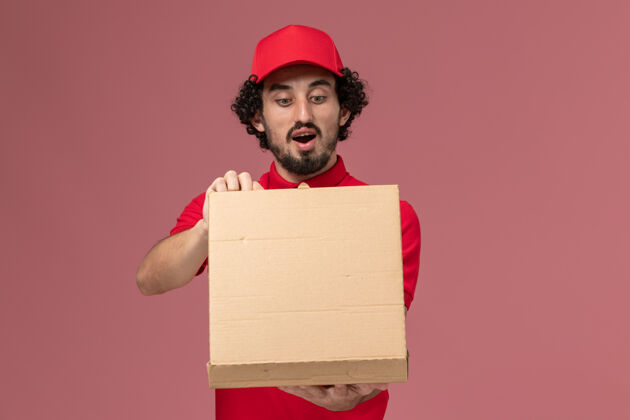 持有正面图身穿红色衬衫和披风的男快递员拿着送货食品箱 在浅粉色的墙上打开正面信封容器