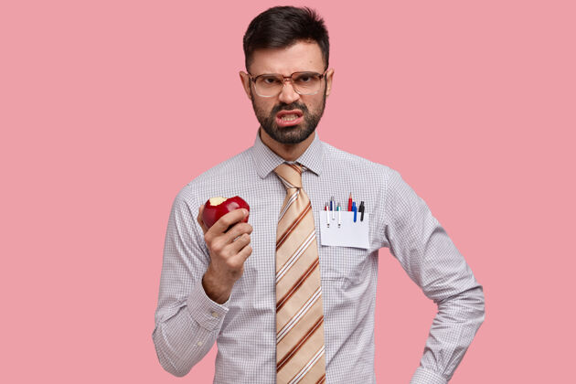 眼镜不满恼怒的胡子男人皱着眉头 气得咬牙切齿 吃着多汁的苹果 不喜欢别人的主意 穿着正装工人人类个人