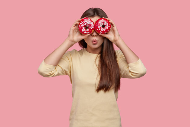 携带美丽小姐的腰上镜头用两个红色的甜甜圈遮住眼睛 穿着休闲服 站在粉色的背景上个性美味美味