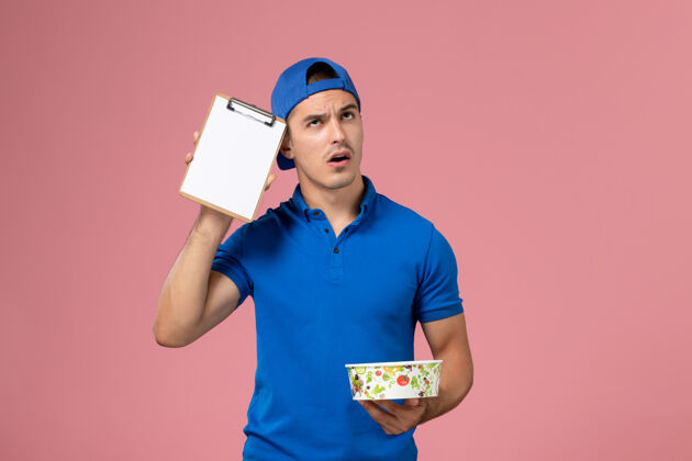 微笑正面图身穿蓝色制服披肩的年轻男性信使拿着记事本和圆形的投递碗在粉红色的墙上思考服务人递送