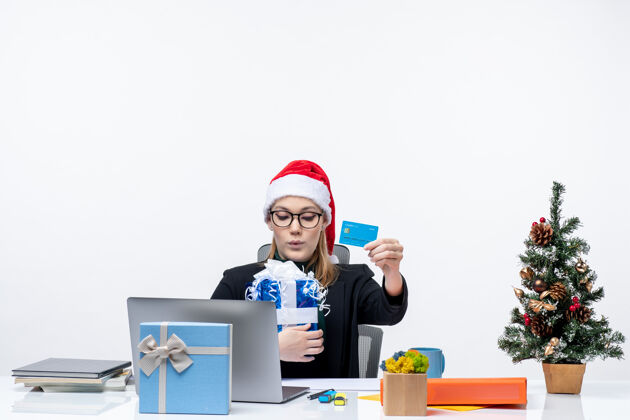桌子戴着圣诞老人帽戴着眼镜的快乐年轻女子坐在桌子旁 手里拿着圣诞礼物和银行卡 背景画面是白色的人笔记本电脑银行